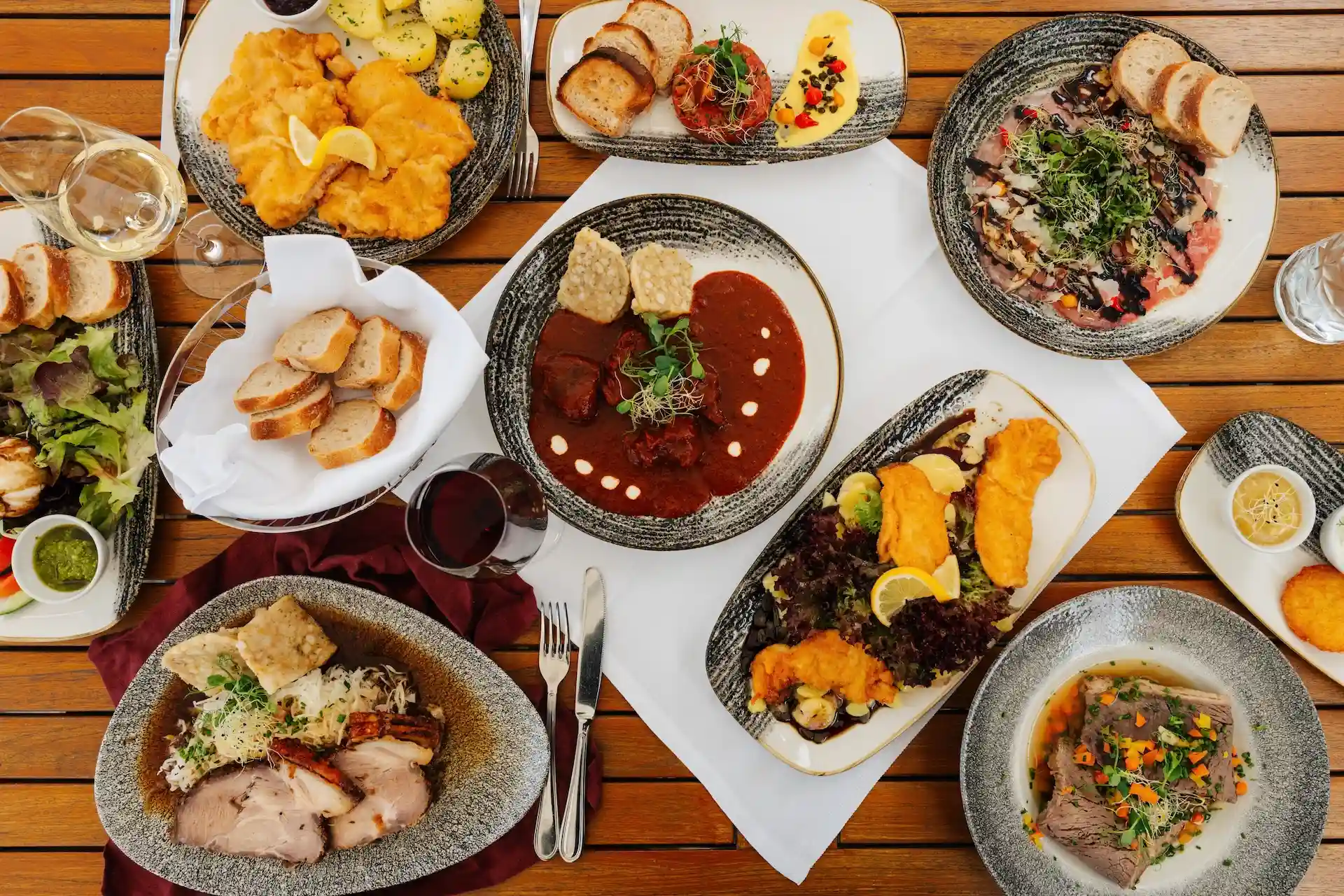 Eine Auswahl an mitteleuropäischen Gerichten, darunter Schnitzel, Gulasch, Braten und Salate, serviert auf mehreren Tellern auf einem Holztisch.