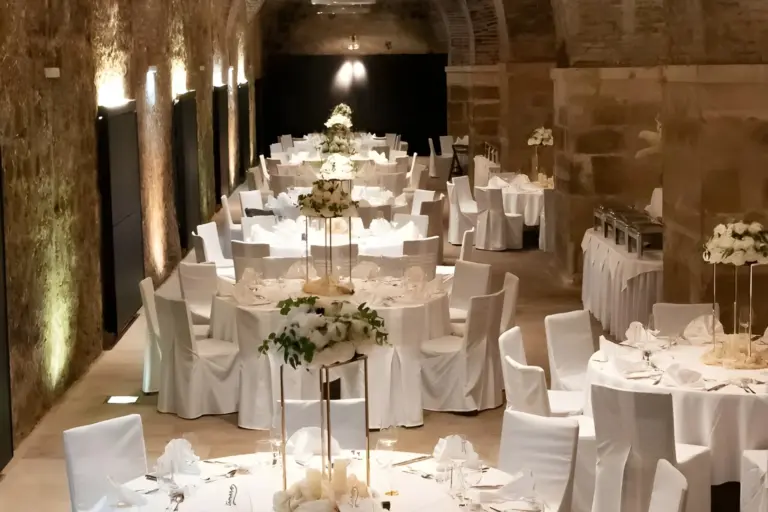 Eleganter Bankettsaal mit Steinwänden und Gewölbedecken, eingerichtet für eine Veranstaltung mit runden, weiß gedeckten Tischen, weißen Stühlen und hohen Blumensträußen. Die sanfte Beleuchtung schafft eine warme, einladende Atmosphäre.
