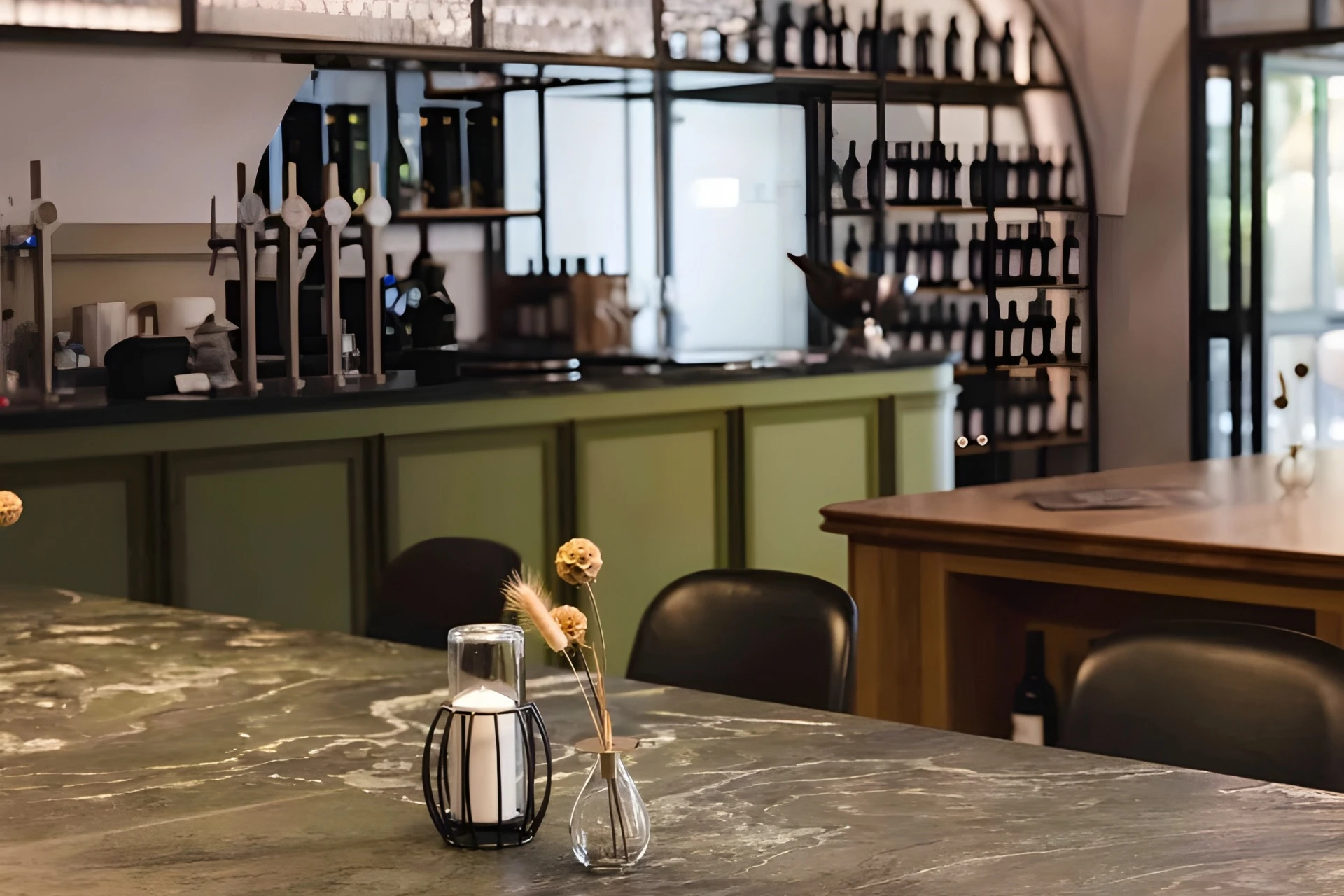 Eine moderne Bar mit einer Arbeitsplatte aus Marmor, schwarzen Stühlen, einer kleinen Vase mit Trockenblumen und einem Kerzenhalter. An der Wand hinter der Bar reihen sich Regale mit Flaschen. Der Raum ist aufgeräumt und sanft beleuchtet.