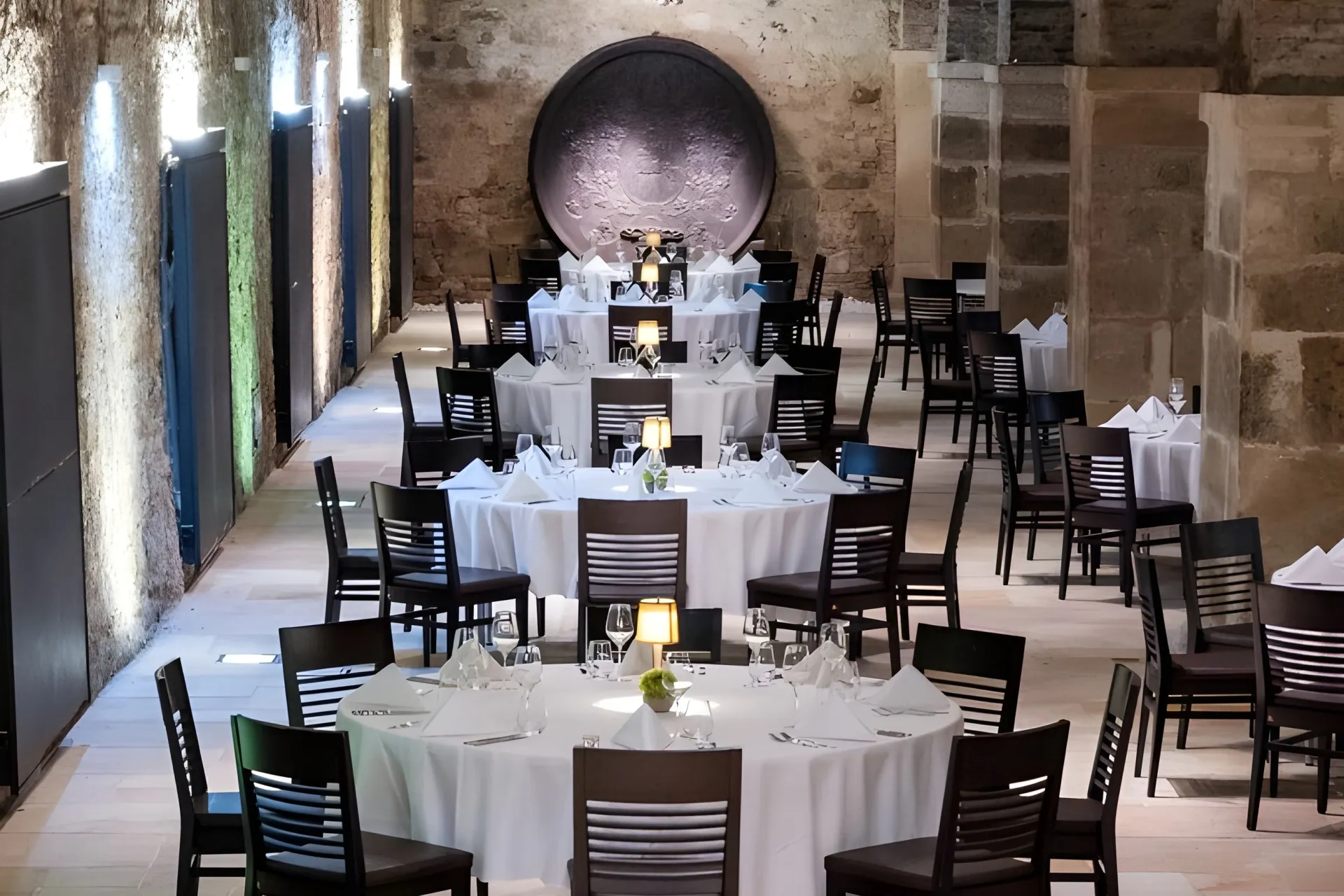 Eleganter Speisesaal des Restaurants mit Steinwänden, runden, weiß gedeckten Tischen, schwarzen Stühlen und sanfter Lampenbeleuchtung, die eine gemütliche und anspruchsvolle Atmosphäre schaffen.