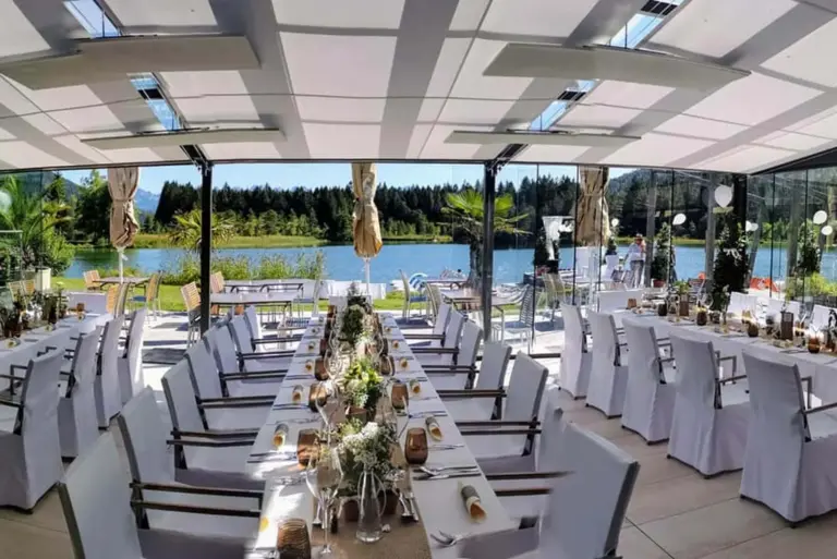Eleganter Hochzeitsempfang mit langen, mit weißen Tüchern bedeckten und mit Blumen und Kerzen geschmückten Tischen, umgeben von Stühlen, in einem hellen Raum mit großen Fenstern und Blick auf einen malerischen See und Bäume.