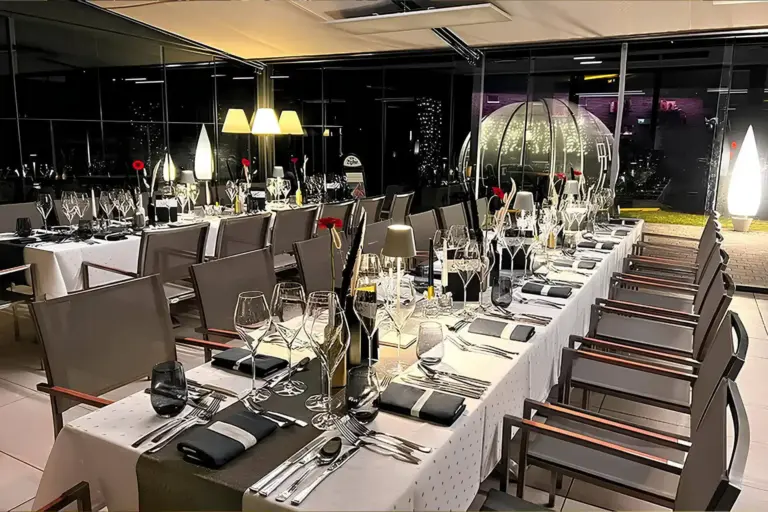 Eleganter Essbereich mit ordentlich gedeckten Tischen, Weingläsern, Silberbesteck, schwarz-weißen Servietten und roten Blumen in der Mitte; große Fenster geben den Blick auf die nächtliche Stadt und die beleuchtete Außendekoration frei.