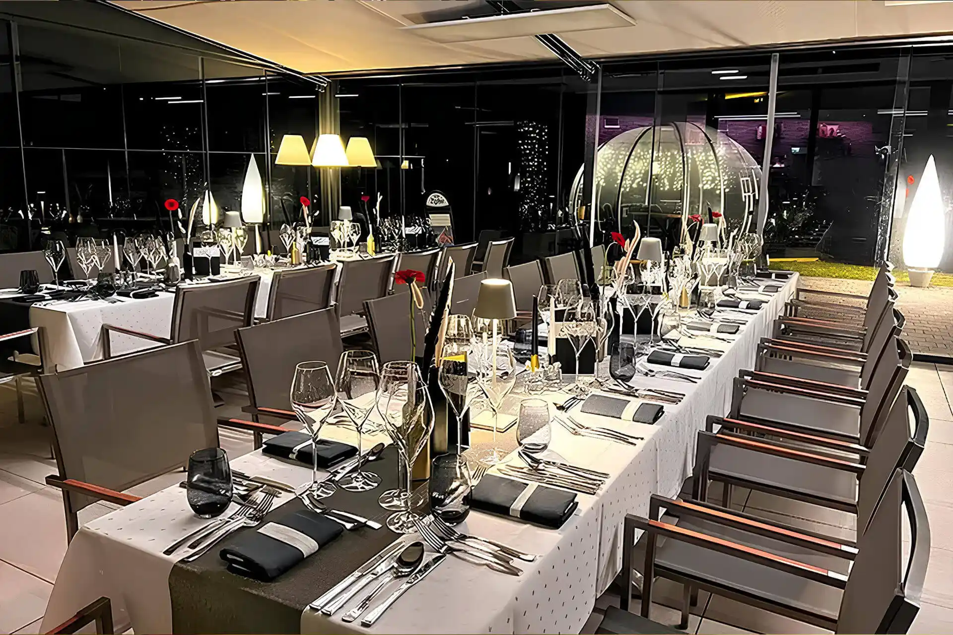 Eleganter Essbereich mit ordentlich gedeckten Tischen, Weingläsern, Silberbesteck, schwarz-weißen Servietten und roten Blumen in der Mitte; große Fenster geben den Blick auf die nächtliche Stadt und die beleuchtete Außendekoration frei.