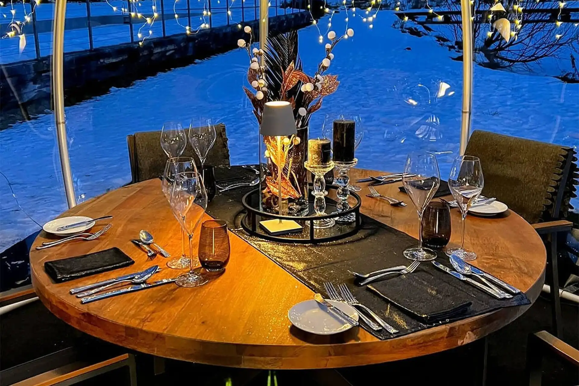 Ein runder Holztisch für fünf Personen mit eleganten Gläsern, Besteck, schwarzen Servietten und dekorativem Tafelaufsatz in einer Kuppel mit Lichterketten und Blick auf eine verschneite Außenlandschaft in der Abenddämmerung.