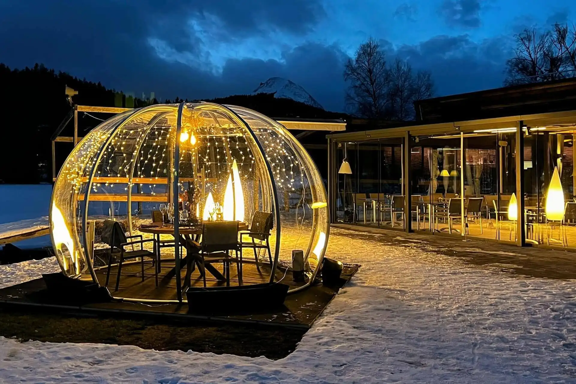 Eine transparente Kuppel mit Lichterketten und einer leuchtenden Lampe im Inneren schützt einen Esstisch und Stühle auf verschneitem Boden neben einem warm beleuchteten Gebäude in der Abenddämmerung, mit Bergen und dunklen Wolken im Hintergrund.