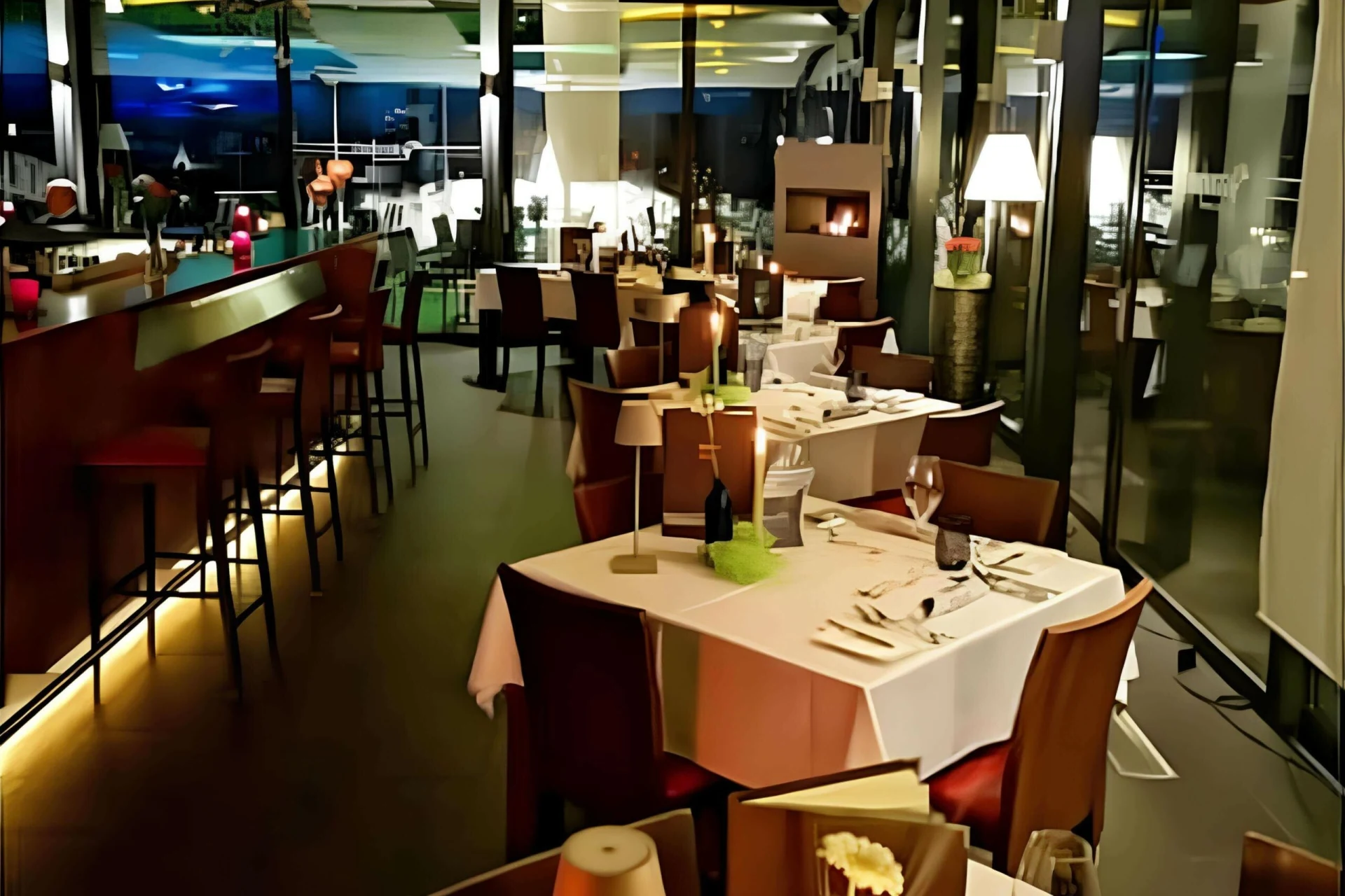 Ein modernes, gehobenes Restaurant mit ordentlich gedeckten Tischen, weißen Tischtüchern, Weingläsern und stimmungsvoller Beleuchtung. Es gibt einen Barbereich mit hohen Hockern und großen Fenstern, die einen nächtlichen Blick auf die Stadt bieten.