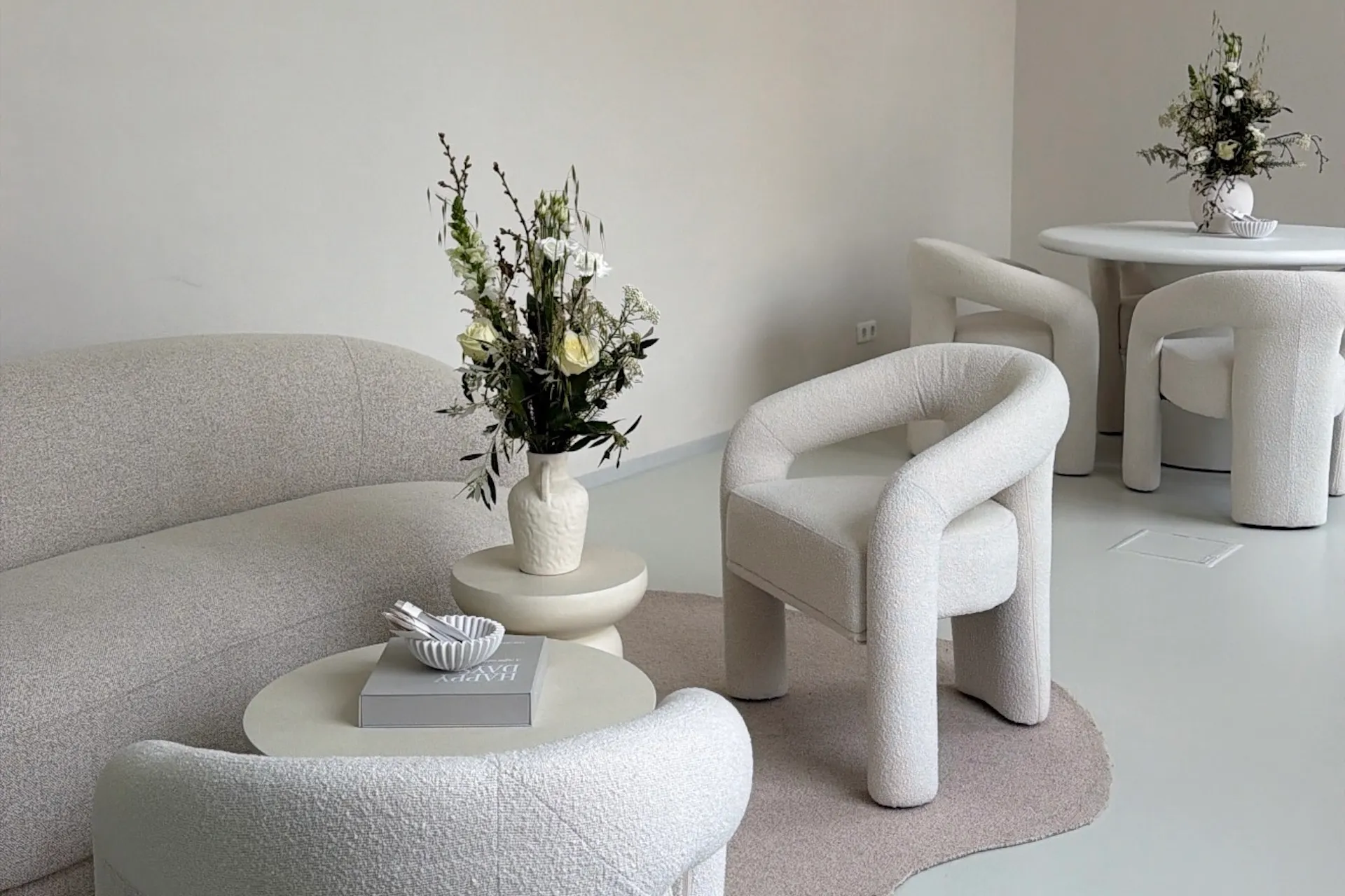 Minimalistisches Wohnzimmer mit einem geschwungenen, cremefarbenen Sofa, Sesseln und runden Tischen. Auf den Tischen stehen Blumenarrangements; die neutrale Einrichtung und das sanfte, natürliche Licht schaffen eine ruhige, elegante Atmosphäre.