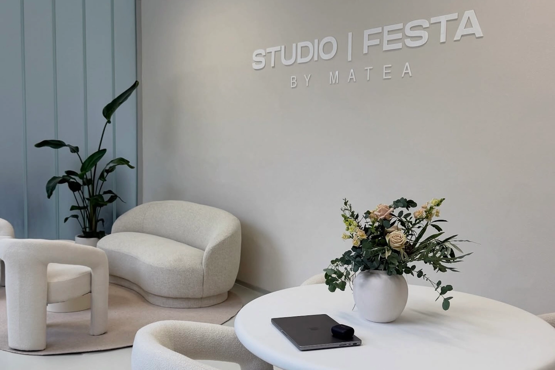 Studio by Matea Minimalistisches Studio mit weißen, modernen Stühlen, einem runden Tisch mit einer Blumenvase, einem Tablet und einem Kameraobjektiv. An der Wand steht „STUDIO I FESTA BY MATEA“. In der Ecke steht eine grüne Pflanze, und der Raum wirkt hell und aufgeräumt.