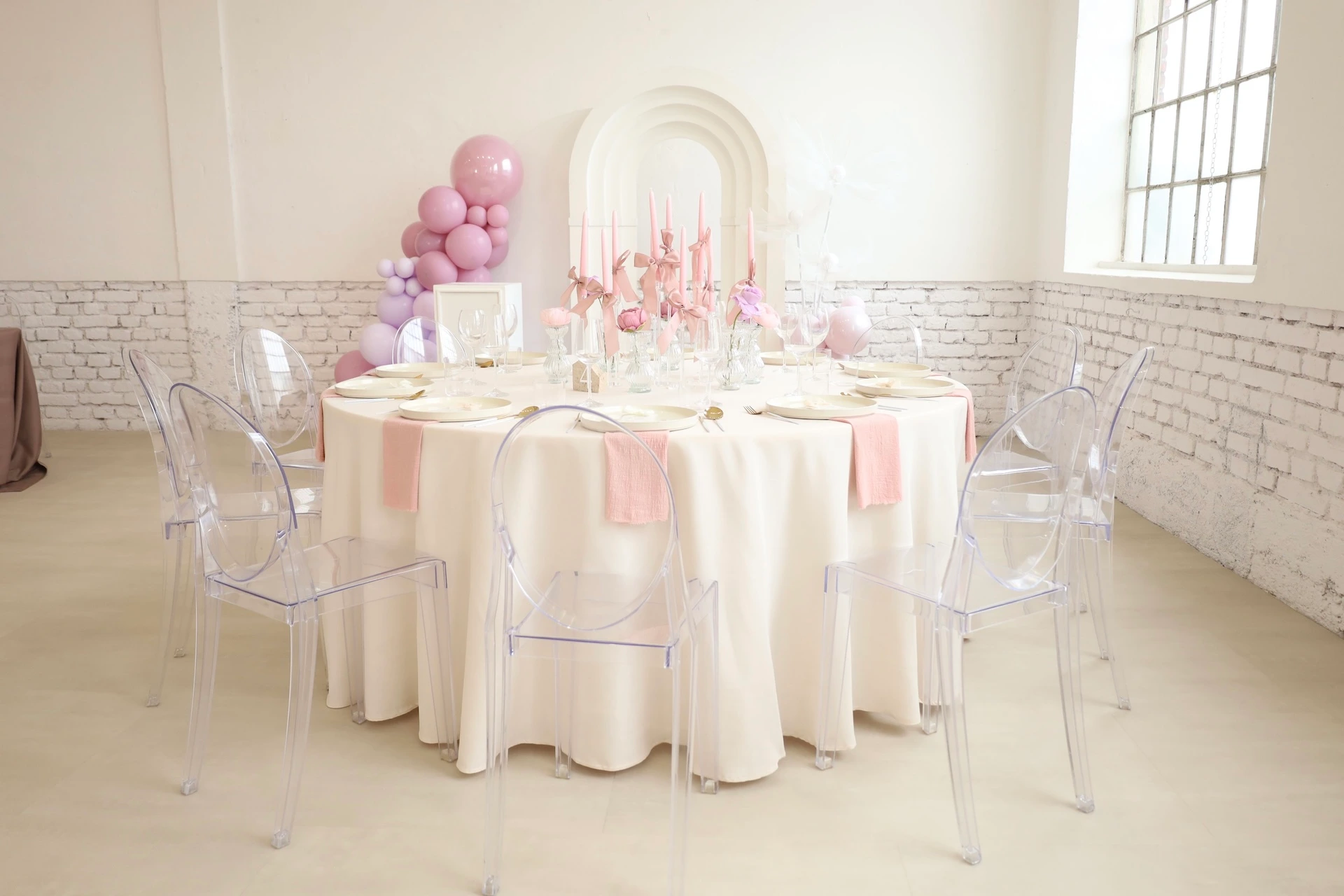 Ein runder Tisch mit cremefarbener Tischdecke ist für sechs Personen gedeckt, mit rosa Servietten, Kerzen und Blumengestecken als Tischdekoration. Transparente Stühle stehen um den Tisch herum, und rosa Luftballons schmücken den Hintergrund. Das Ambiente ist elegant und luftig.