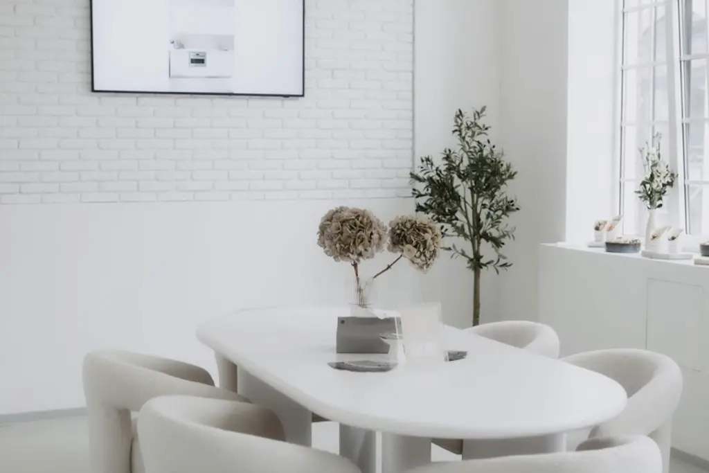 Minimalistisches weißes Esszimmer mit einem rechteckigen Tisch, sechs gepolsterten Stühlen, getrockneten Hortensien in einer Vase, einer Topfpflanze, einem großen Fenster und einer Akzentwand aus Ziegelsteinen, über der ein Fernseher oder ein gerahmtes Bild hängt.