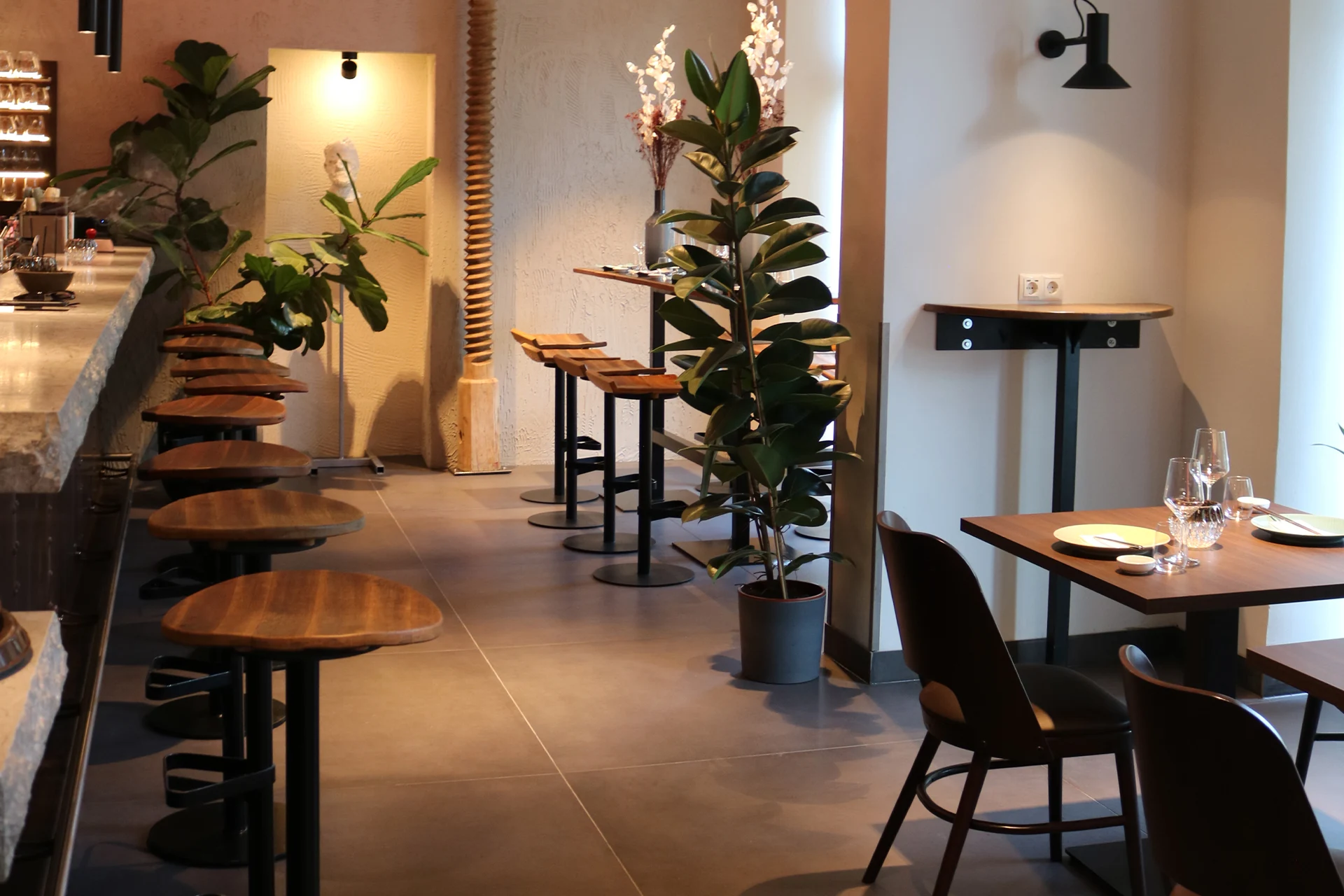 Ein modernes Restaurant mit Holztischen und -stühlen, Topfpflanzen, gedämpfter Beleuchtung und Gedecken auf den Tischen. Die Atmosphäre wirkt ruhig und einladend.