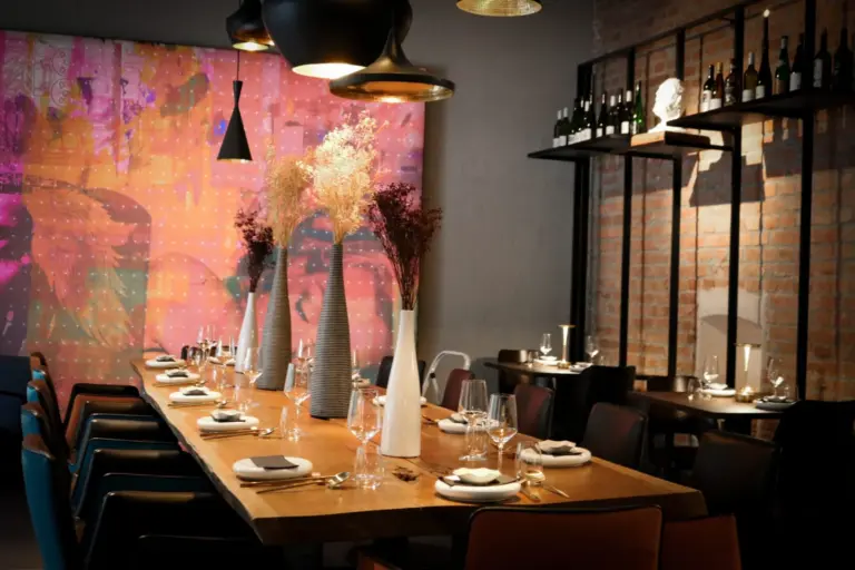 Ein modernes Restaurant mit einem langen Holztisch zum Essen, modernen Pendelleuchten, Vasen mit Trockenblumen, freiliegenden Ziegelwänden und bunter abstrakter Kunst an einer Wand. Weinflaschen sind in Regalen ausgestellt.