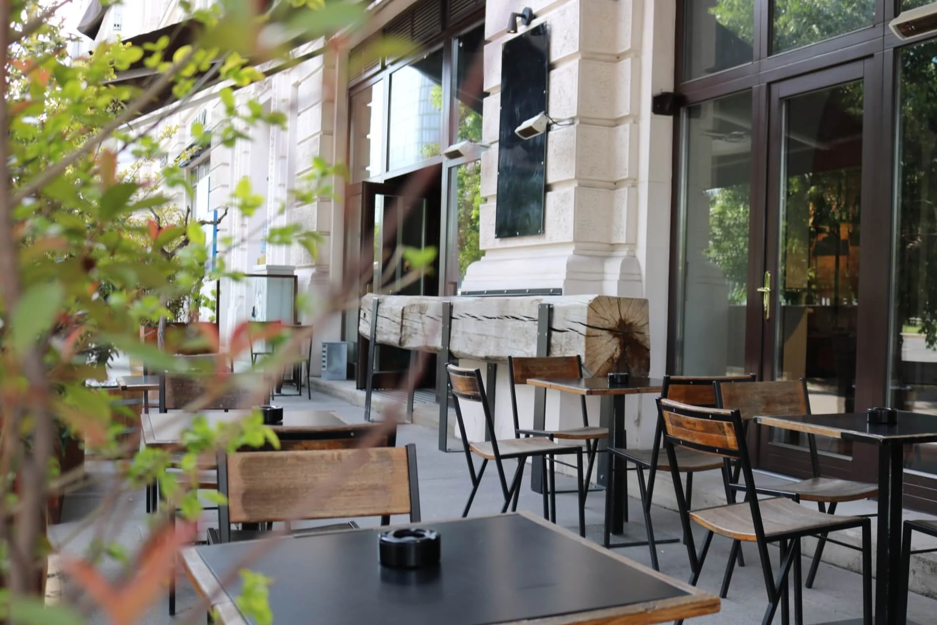 Außencafé mit leeren Holztischen und -stühlen, die entlang eines Gehwegs neben einem Gebäude mit großen Fenstern angeordnet sind; im Vordergrund sind grüne Blattpflanzen zu sehen.