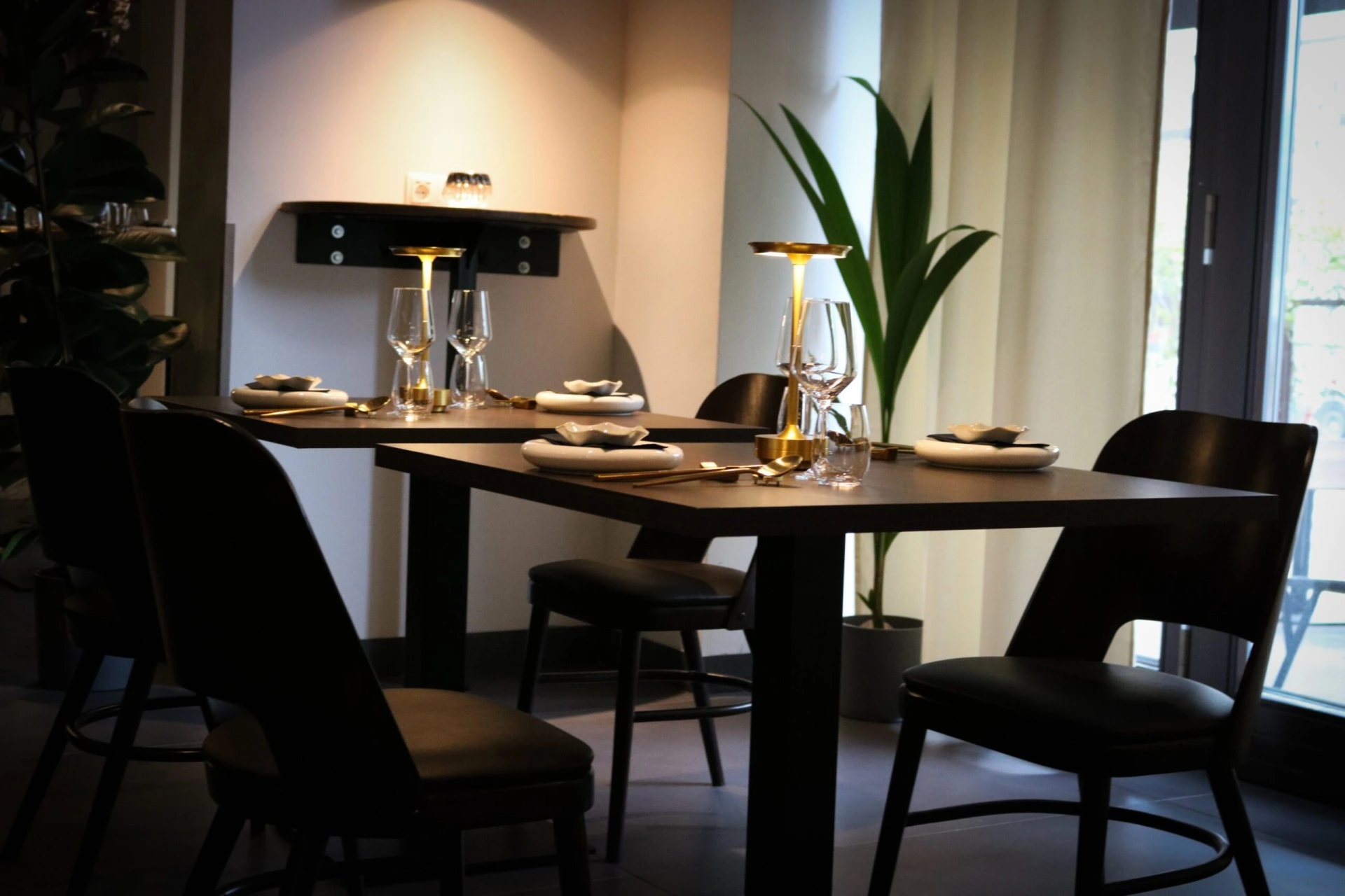 Ein moderner Restauranttisch für vier Personen mit schwarzen Stühlen, weißen Tellern, gefalteten Servietten, Weingläsern und goldenen Lampen, neben hohen Pflanzen und großen Fenstern mit transparenten Vorhängen.