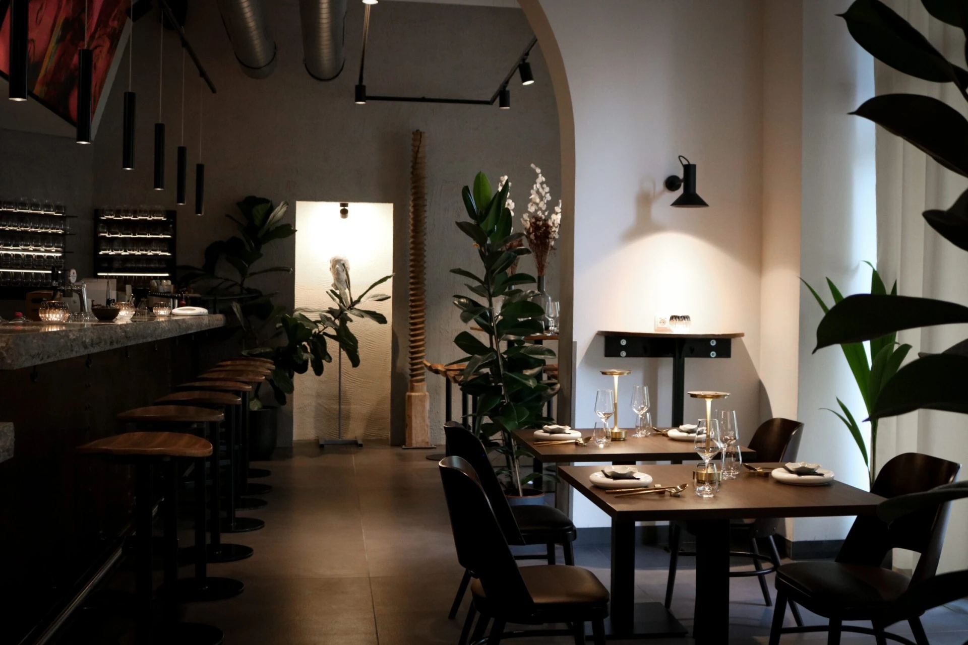 Ein modernes, schwach beleuchtetes Restaurant mit Holztischen zum Essen, schwarzen Stühlen, einer langen Bar mit Hockern, grünen Topfpflanzen und minimalistischer Einrichtung. Durch ein Fenster fällt sanftes natürliches Licht, das eine gemütliche Atmosphäre schafft.