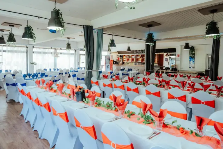Ein Bankettsaal mit langen Tischen, die mit weißen Tüchern bedeckt und mit roten und blauen Stuhlschärpen, roten Servietten, Grünzeug und Blumen geschmückt sind, gedeckt für eine große Veranstaltung, mit einer Bar im Hintergrund.