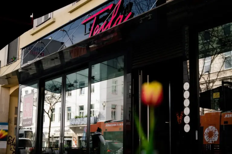 Ein gläsernes Gebäude mit der Leuchtreklame Tesla über dem Eingang, in der sich die umliegenden Gebäude und Bäume spiegeln. Eine einzelne rote und gelbe Tulpe erscheint unscharf im Vordergrund.