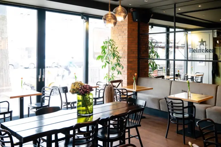 Ein helles, modernes Café mit Holztischen und schwarzen Stühlen, einer großen Blumenvase auf einem Tisch, Pendelleuchten, Pflanzen und einem Heineken-Schild an einer verspiegelten Wand. Sonnenlicht strömt durch große Fenster.