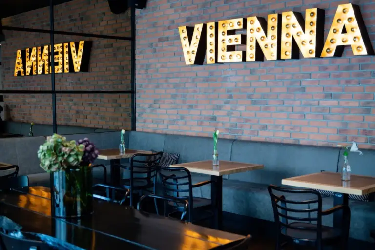 Ein modernes Café mit Backsteinwänden, kleinen Tischen, schwarzen Stühlen und dem Schriftzug WIEN, der auf einer großen Markise prangt. Das Wort wird auch in einem Spiegel an der linken Wand reflektiert. Vasen mit einzelnen Blumen schmücken die Tische.