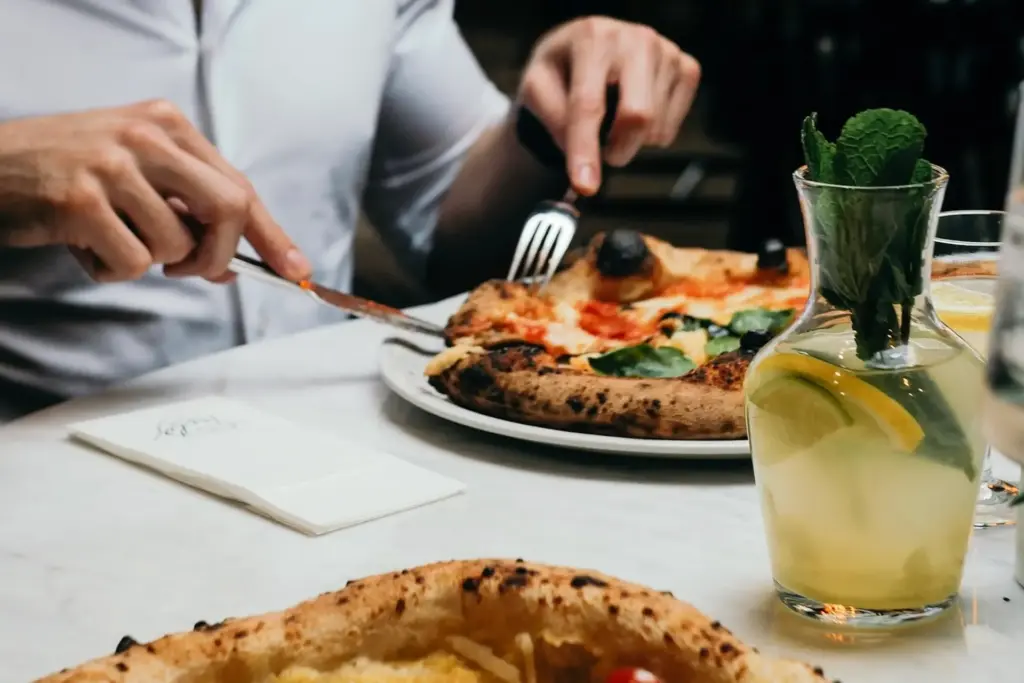 Eine Person in einem weißen Hemd schneidet an einem Tisch mit Messer und Gabel eine Pizza an. Neben der Pizza steht ein Glaskrug mit Limonade, Zitronenscheiben und Minze. Eine weitere Pizza ist teilweise im Vordergrund zu sehen.