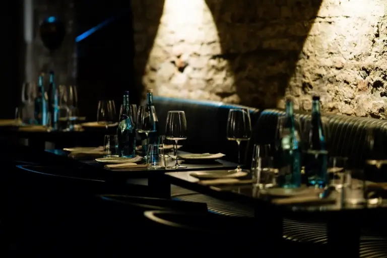Ein schwach beleuchtetes Restaurant mit gedeckten Tischen, auf denen leere Gläser, Teller und blaue Wasserflaschen auf schwarzen Tischen vor einer Backsteinwand stehen.