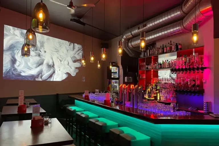 Eine moderne Bar mit Neonbeleuchtung, Reihen von Flaschen in Regalen, Barhockern und Hängelampen. Ein großes abstraktes Bild wird an die Wand projiziert, und die Atmosphäre ist bunt und stilvoll.
