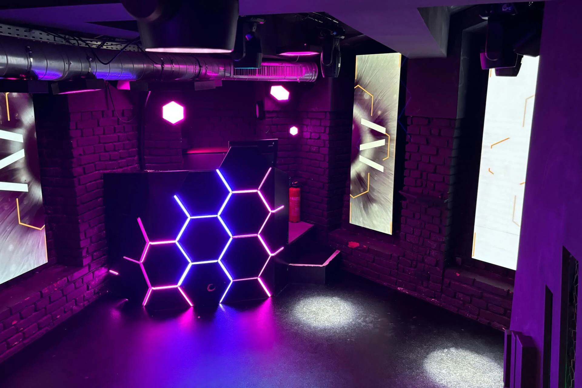Ein schummrig beleuchteter Nachtclub mit einem DJ-Pult mit sechseckigen Neonröhren in Lila und Rosa, freiliegenden Backsteinwänden und großen digitalen Bildschirmen, die abstrakte Bilder zeigen.