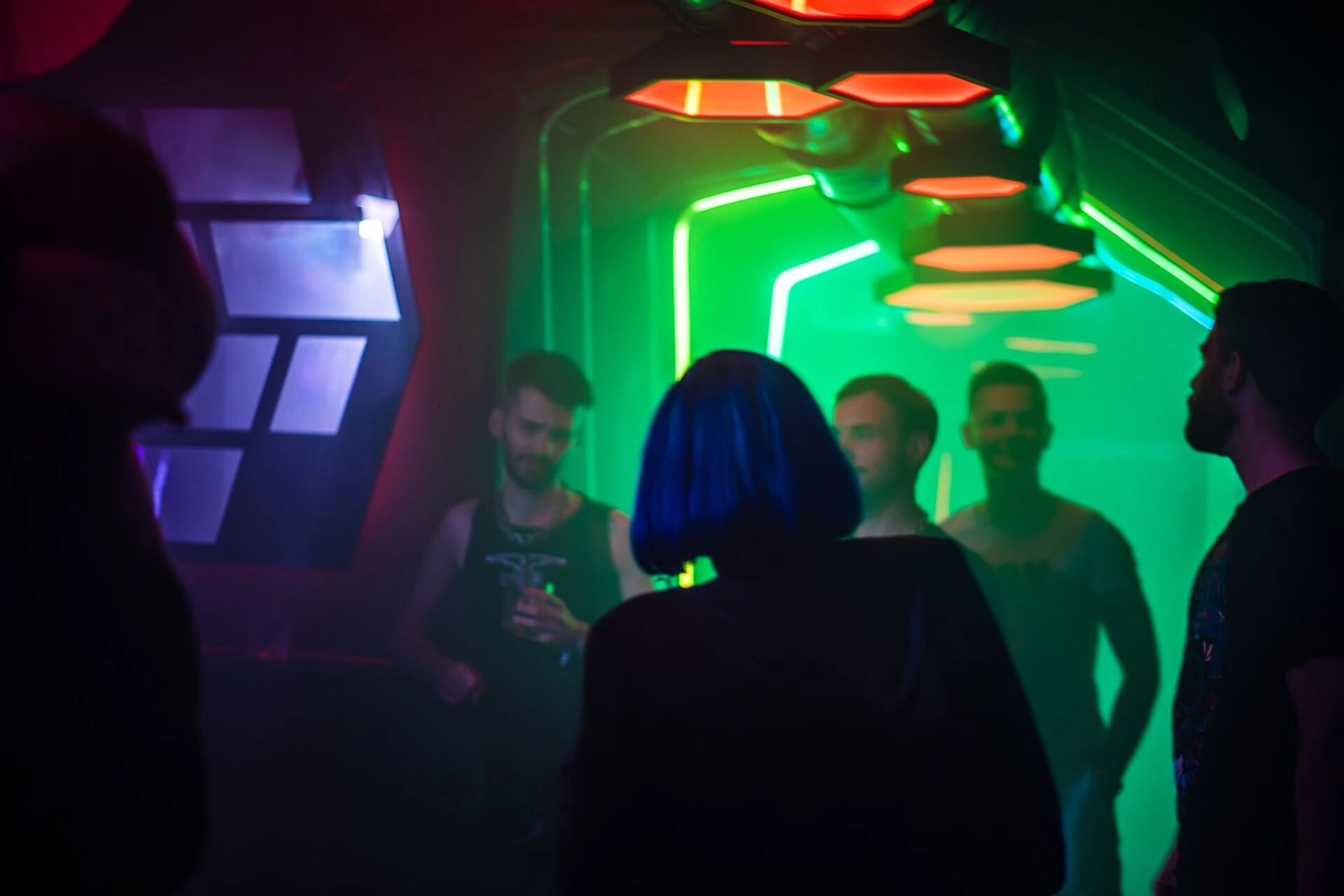 Menschen stehen in einem schwach beleuchteten Club mit neongrünen und roten Lichtern. Eine Person mit kurzen blauen Haaren steht im Vordergrund und blickt auf eine Gruppe von Männern, die sich unterhalten und Getränke in der Hand halten. Die Atmosphäre ist lebhaft und futuristisch.