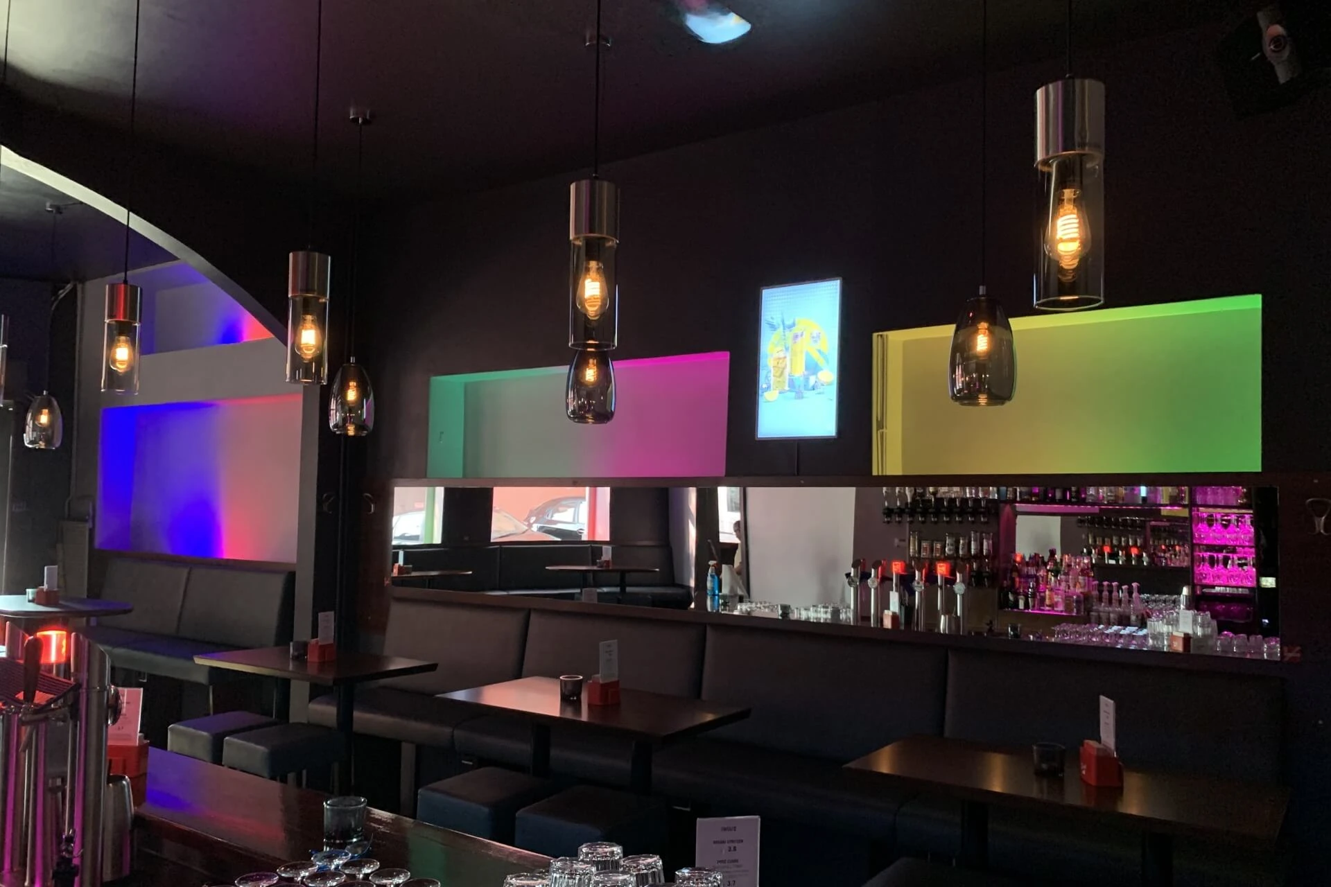 Eine moderne Bar mit hängenden Pendelleuchten, dunklen Möbeln und einem langen Spiegel hinter der Bar. Sanfte farbige Lichter in Lila, Rosa, Grün und Gelb beleuchten Teile der Rückwand.