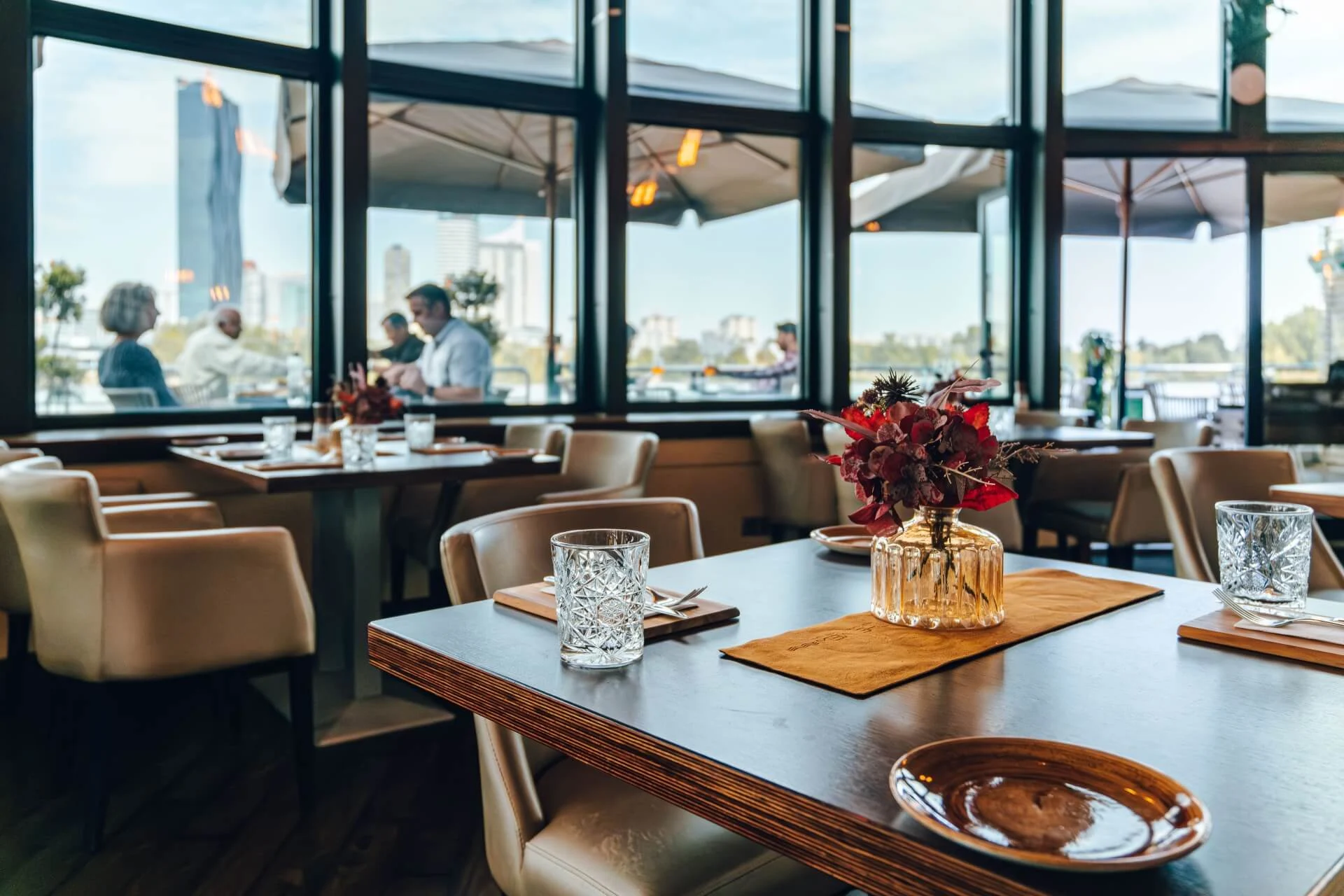 Ein modernes Restaurant mit elegant gedeckten Tischen, Glastassen und einer Vase mit Trockenblumen. Große Fenster geben den Blick auf eine Terrasse frei, auf der Menschen sitzen, und im Hintergrund sind städtische Gebäude zu sehen.