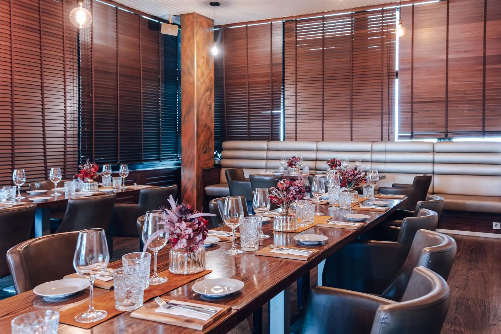 Eleganter Essbereich im Restaurant mit langen Holztischen, die für eine Mahlzeit gedeckt sind, mit Gläsern, Tellern, Servietten und Blumenschmuck. Braune Lederstühle und Holzjalousien schaffen eine warme, moderne Atmosphäre.