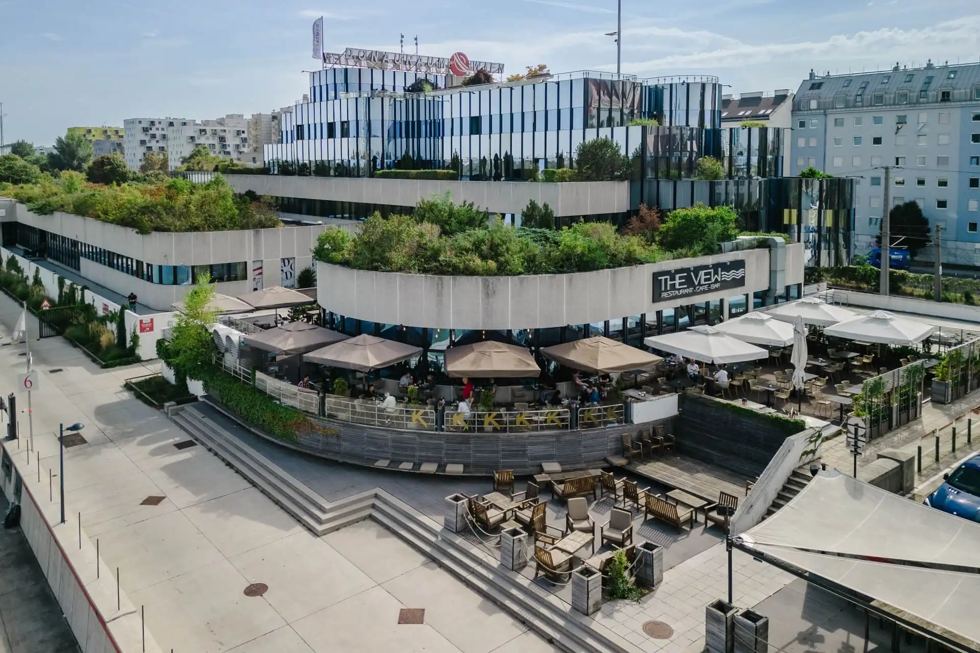Ein modernes Gebäude mit Dachgärten und Terrassen, das ein Außenrestaurant mit Tischen, Sonnenschirmen und Sitzgelegenheiten bietet. In der Umgebung gibt es breite Stufen und einen großen, offenen Platz.
