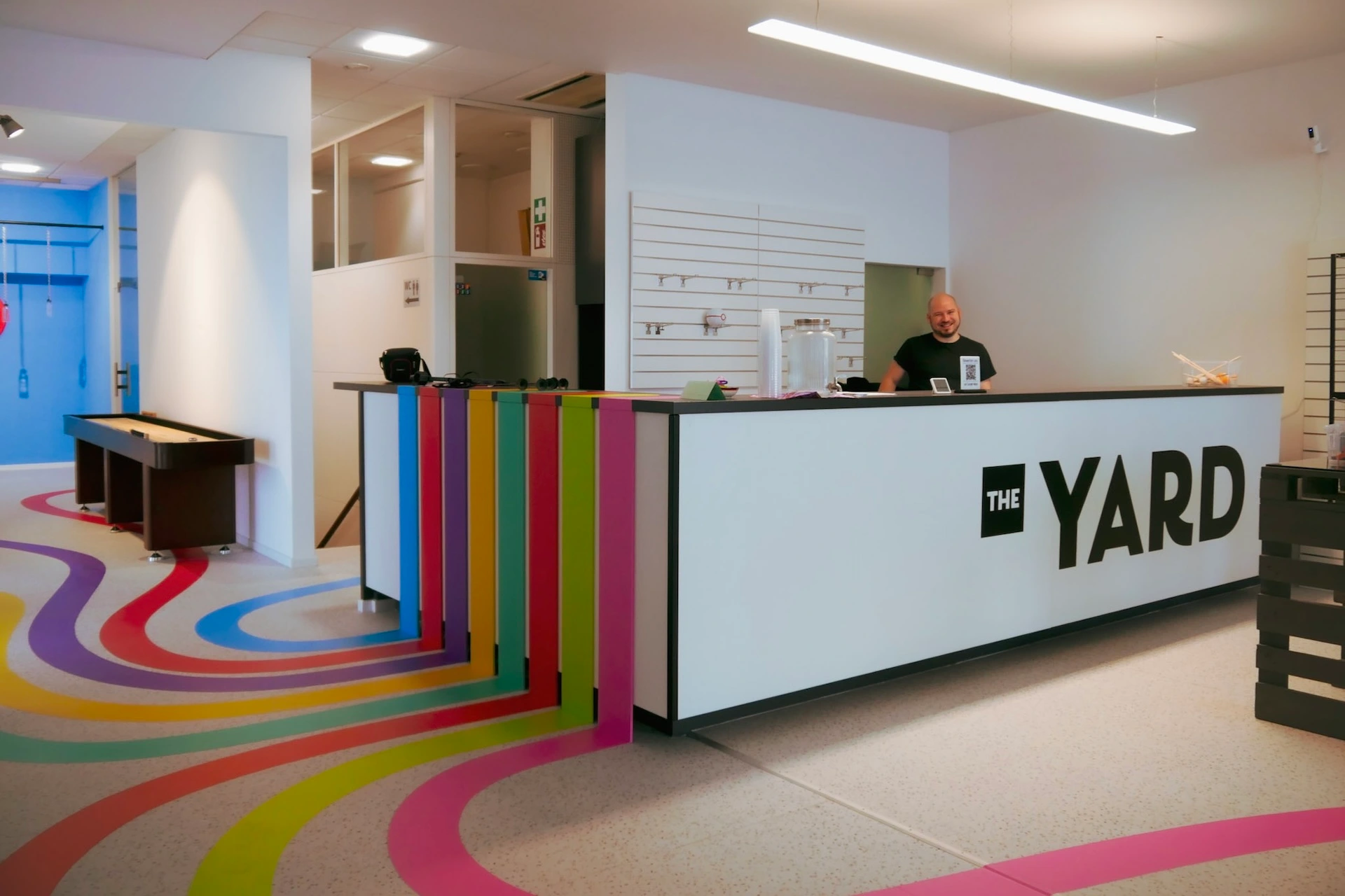 Ein moderner Empfangsbereich mit einer weißen Theke mit der Aufschrift THE YARD. Bunte Streifen auf dem Boden ziehen sich bis zum Tresen hinauf. Eine lächelnde Person steht hinter dem Tresen. Der Raum ist sauber, hell und modern.