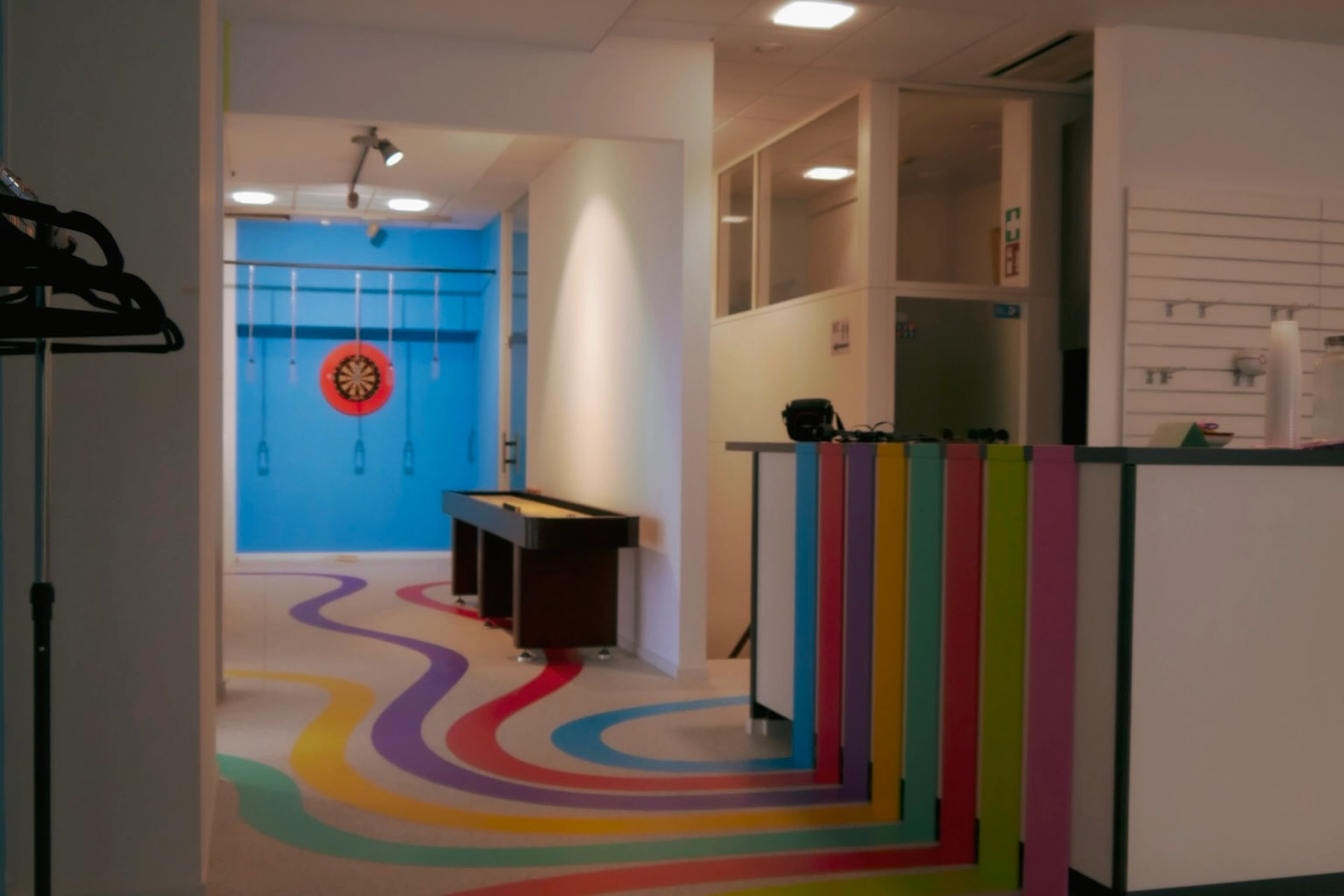 THE YARD Ein farbenfroher Büroraum mit wellenförmigen Streifen auf dem Boden und dem Schreibtisch, die zu einer blauen Wand mit einer Dartscheibe und Gymnastikringen führen. Auch Garderobenständer und moderne Beleuchtung sind zu sehen.