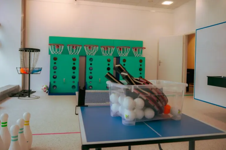 Ein Fitnessraum mit türkisfarbenen Spinden, Basketballkörben, einem Discgolf-Korb, Bowlingkegeln und einem Tischtennistisch mit einem durchsichtigen Behälter, der mit Schlägern und weißen Tischtennisbällen gefüllt ist.