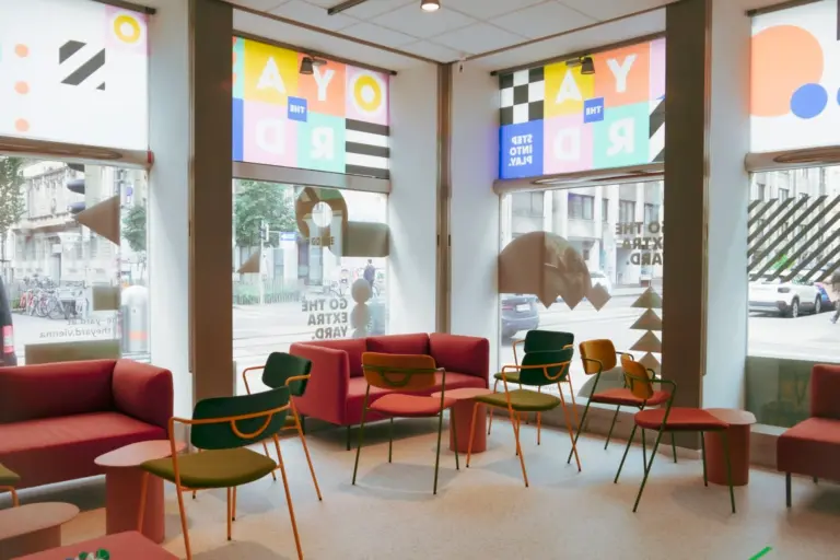 THE YARD Ein modernes Wartezimmer mit bunten Stühlen, rosa Sofas, kleinen Tischen und großen Fenstern, die mit hellen geometrischen Mustern und motivierenden Texten verziert sind. Die Straße und Gebäude der Stadt sind von außen zu sehen.