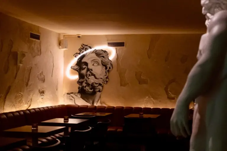 Ein schummriges Wohnzimmer mit braunen Ledersesseln und leeren Tischen. Eine große Wandskulptur eines bärtigen Gesichts mit einem haloartigen Licht befindet sich an der Rückwand, und im Vordergrund ist ein Teil einer weißen Statue zu sehen.