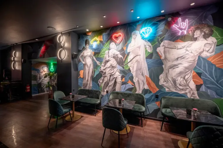 toga Eine stilvolle Lounge mit Samtstühlen und Marmortischen, die mit einer lebhaften Wandmalerei klassischer Statuen und Neonschildern in Form eines Herzens, einer Raute und einer Handgeste geschmückt ist.