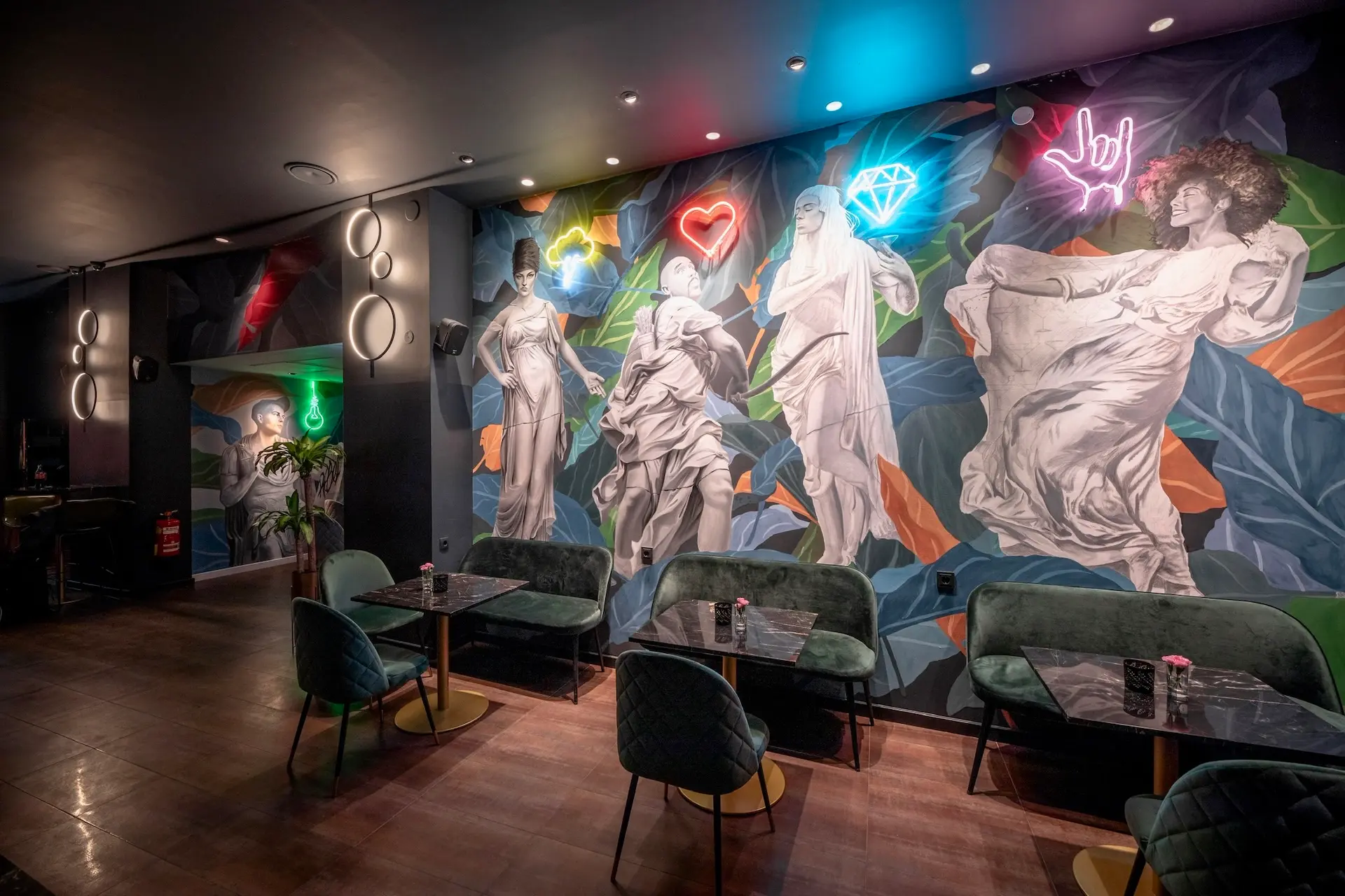 toga Eine stilvolle Lounge mit Samtstühlen und Marmortischen, die mit einer lebhaften Wandmalerei klassischer Statuen und Neonschildern in Form eines Herzens, einer Raute und einer Handgeste geschmückt ist.