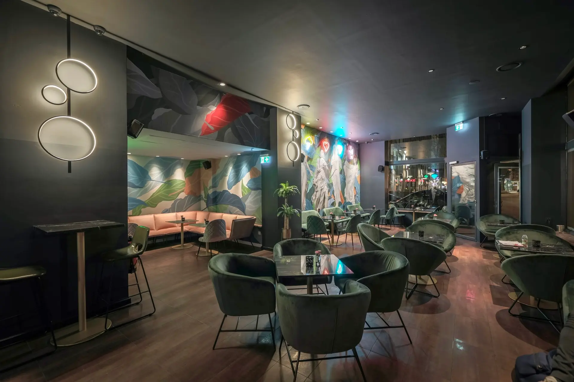 Eine moderne Lounge-Bar mit grünen Samtstühlen, Glastischen, zartrosa Sofas und bunter Wandkunst. Die Beleuchtung ist gedämpft und stilvoll und schafft eine gemütliche Atmosphäre mit dekorativen Wandleuchten und Pflanzen.