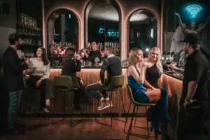 In einer stilvollen Bar mit schummriger Beleuchtung kommen die Menschen zusammen. Einige sitzen auf Barhockern, andere stehen, plaudern und trinken. An der Wand hinter dem Tresen leuchtet ein Neonschild im Toga Wien.