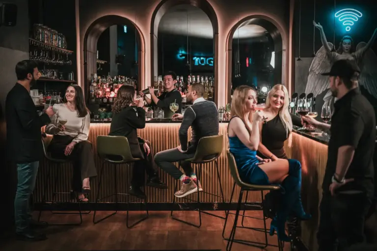 In einer stilvollen Bar mit schummriger Beleuchtung kommen die Menschen zusammen. Einige sitzen auf Barhockern, andere stehen, plaudern und trinken. An der Wand hinter dem Tresen leuchtet ein Neonschild mit einem Wi-Fi-Engel.