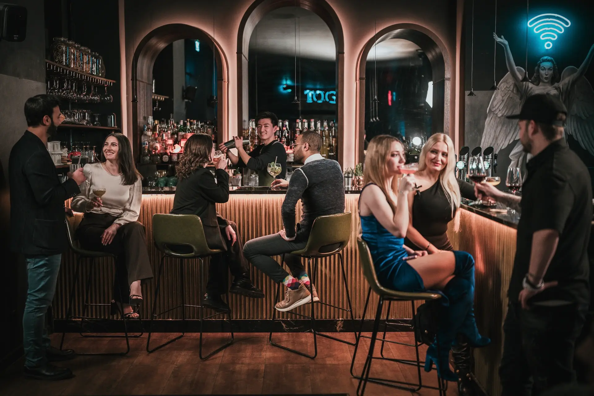 In einer stilvollen Bar mit schummriger Beleuchtung kommen die Menschen zusammen. Einige sitzen auf Barhockern, andere stehen, plaudern und trinken. An der Wand hinter dem Tresen leuchtet ein Neonschild mit einem Wi-Fi-Engel.
