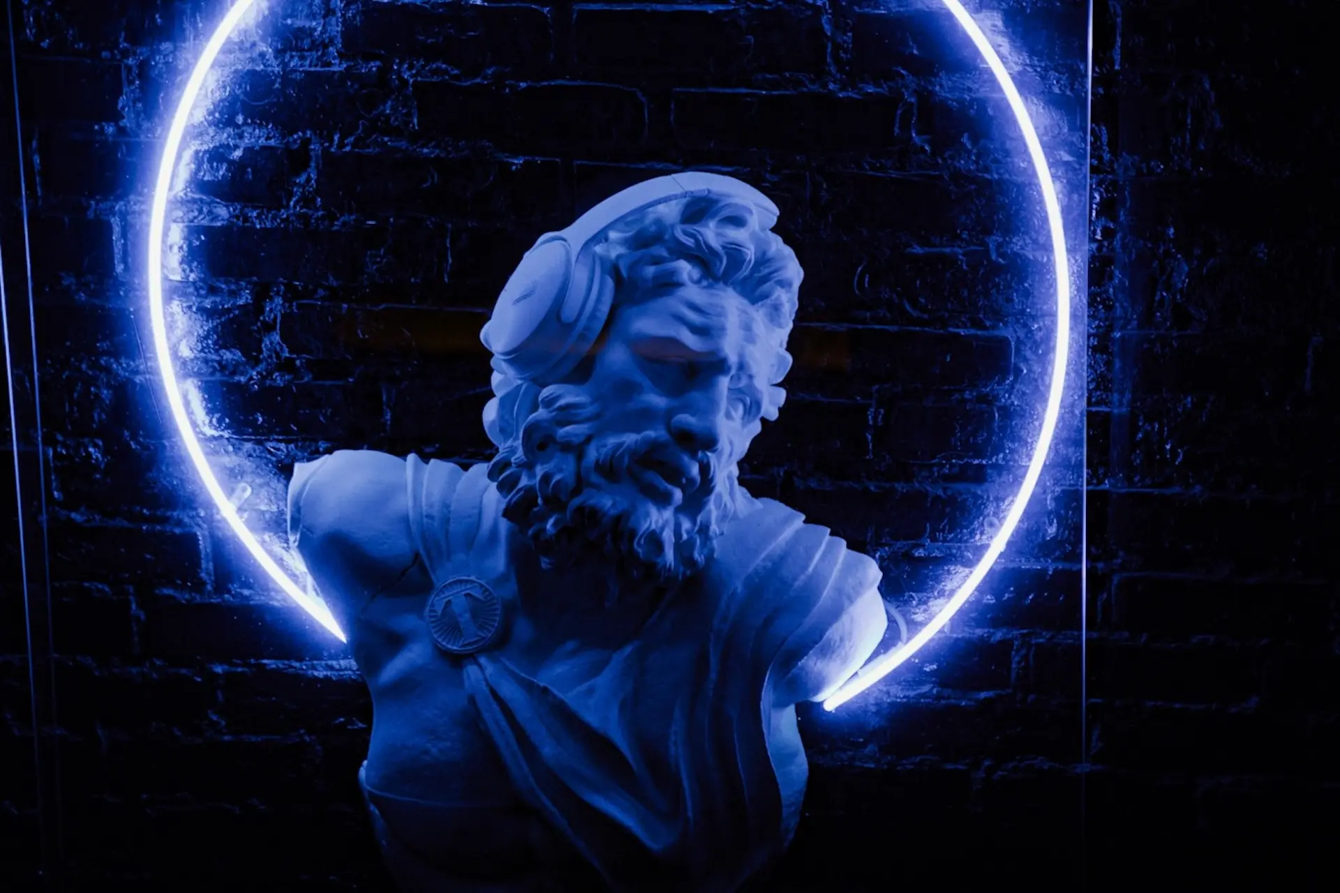 Eine blau beleuchtete, klassizistische Statue mit Kopfhörern steht vor einer dunklen Backsteinmauer, eingerahmt von einem leuchtenden neonblauen Kreis.