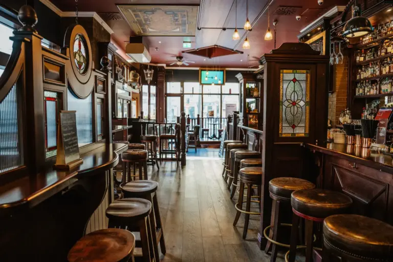 Eine leere Kneipe mit Holzhockern, hohen Tischen, einer langen Bar, dekorativen Buntglasscheiben und mit Flaschen gefüllten Regalen, warm beleuchtet von Hängelampen und natürlichem Licht aus großen Fenstern.