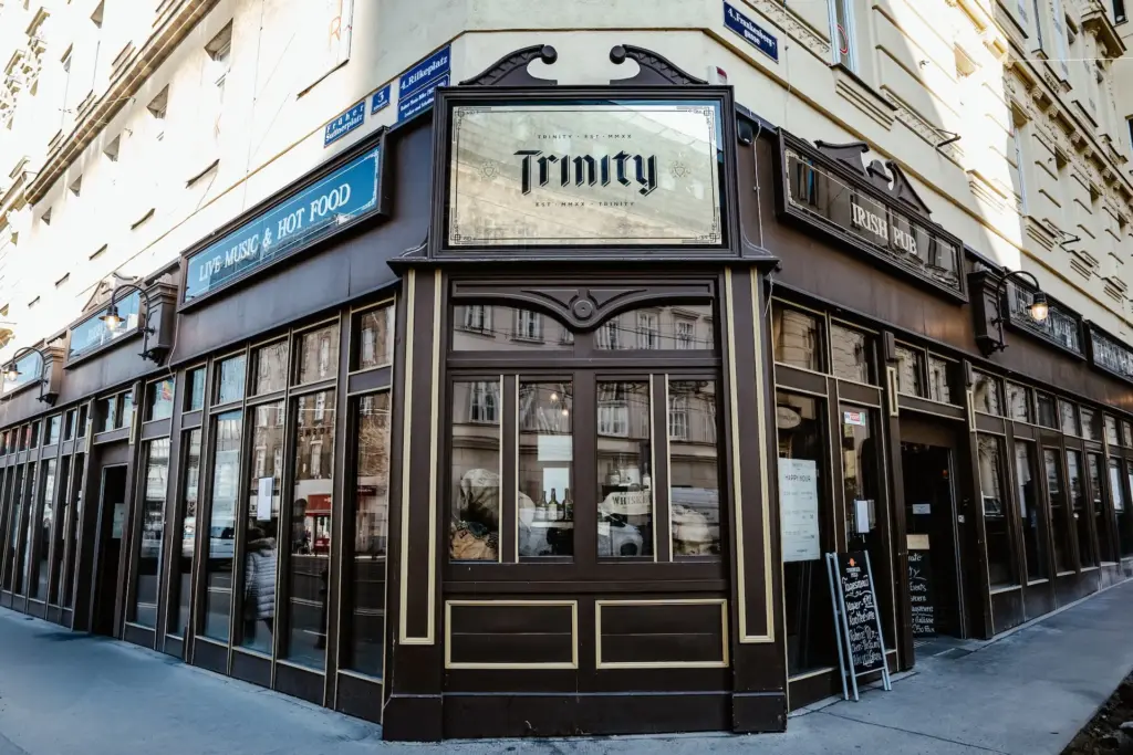 Eckansicht eines traditionellen Pubs mit dem Namen "Trinity" mit großen Fenstern, braunen Zierleisten, alten Schildern und Speisekarten auf Kreidetafeln im Freien an einer Stadtstraße. Das Äußere des Gebäudes ist cremefarben und mit architektonischen Details verziert.