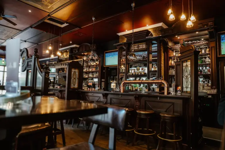 Eine gemütliche Kneipeneinrichtung mit dunklen Holzmöbeln, einer langen Bar mit Hockern, mit Flaschen gefüllten Regalen, Hängelampen und Flachbildfernsehern über der Bar. Der Raum ist warm beleuchtet und einladend.