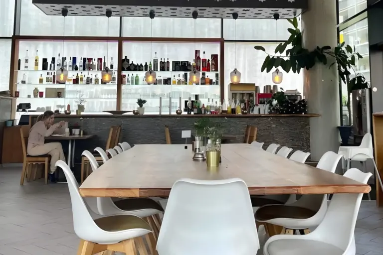 Modernes Café mit einem großen Holztisch und weißen Stühlen im Vordergrund, Regalen mit Flaschen hinter der Theke, Topfpflanzen und zwei Personen, die an einem Beistelltisch sitzen. Helle, natürliche Beleuchtung erfüllt den Raum.