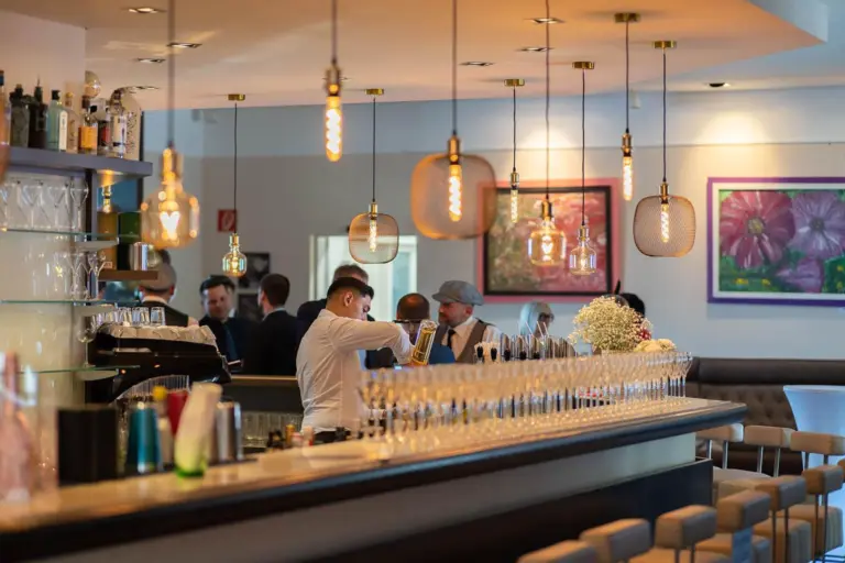 Ein Barkeeper bereitet hinter einer modernen, mit leeren Gläsern bestückten Bar Getränke zu, während sich im Hintergrund mehrere Personen unterhalten. Warme Hängelampen und modernes Dekor schaffen eine gemütliche, gehobene Atmosphäre.