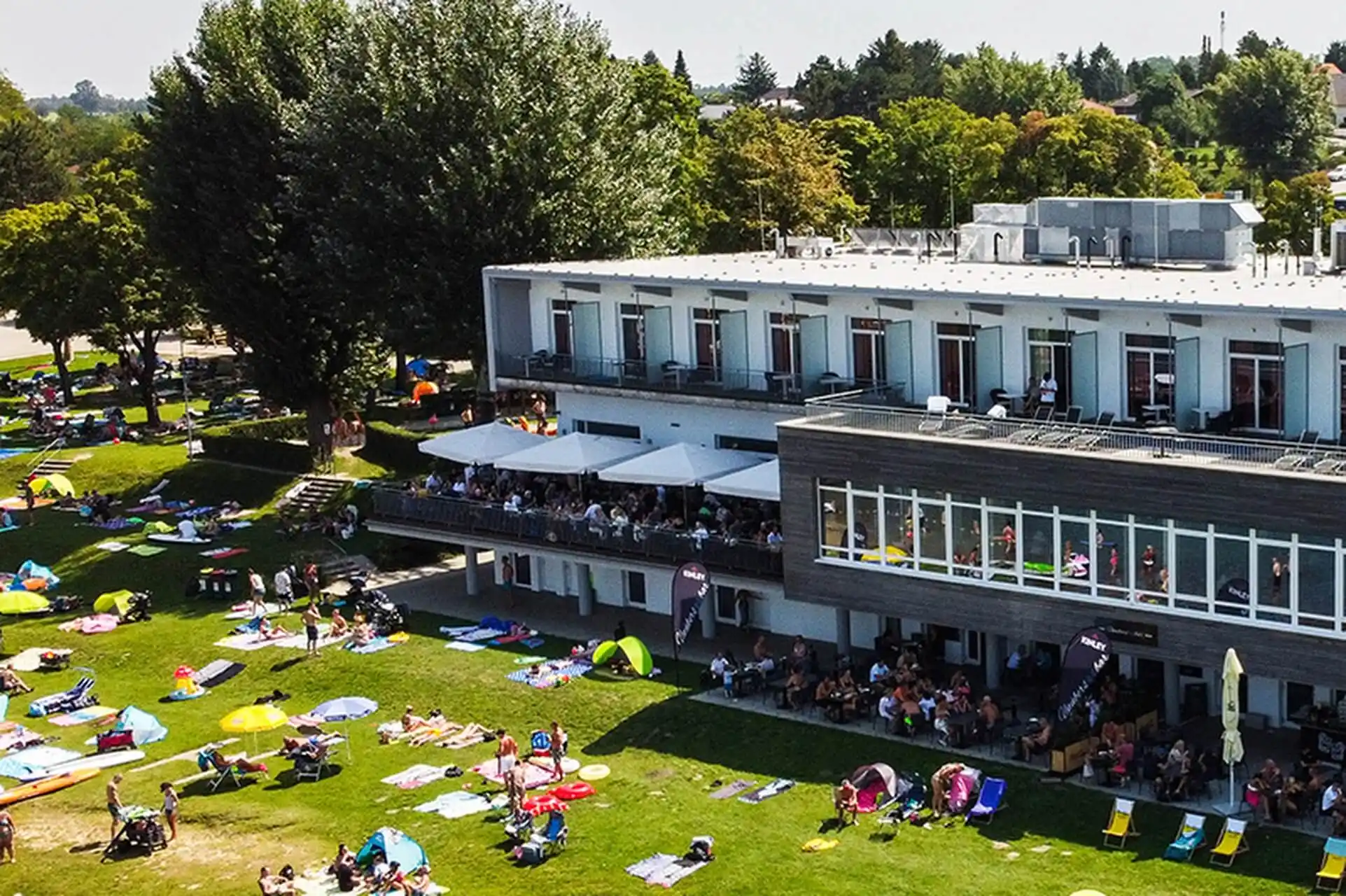 Unüberseebar Ein modernes zweistöckiges Gebäude blickt auf eine Rasenfläche, auf der sich die Leute auf Handtüchern und unter Sonnenschirmen entspannen. Hier herrscht reges Treiben, Gruppen genießen den sonnigen Tag in der Nähe von Bäumen und Grünanlagen.